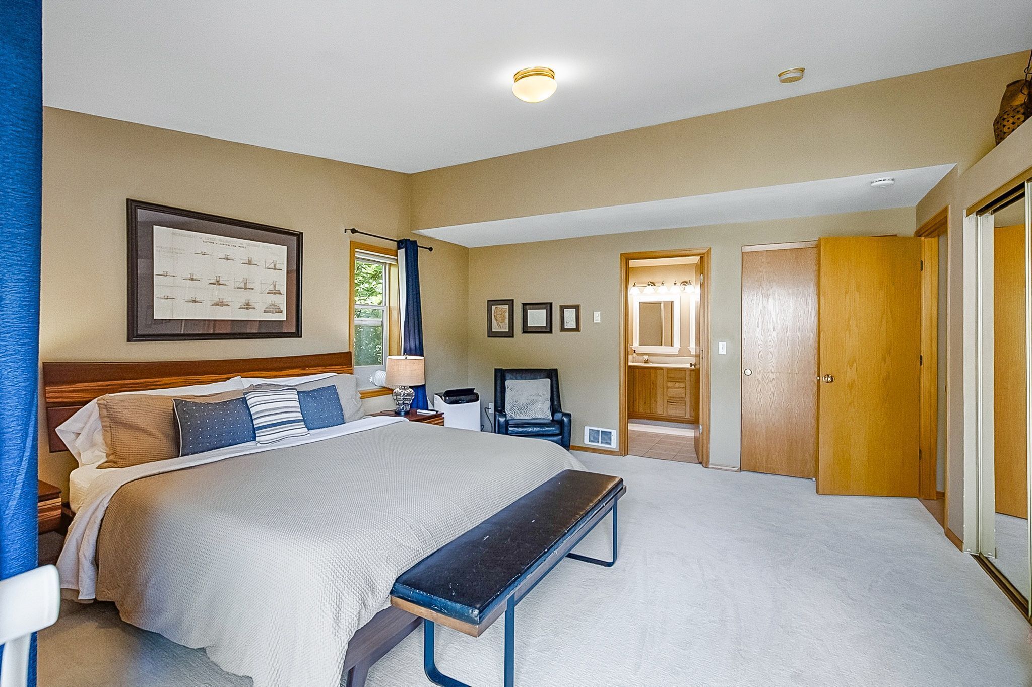 A bedroom with a large bed, a bench, a chair, a desk, and a framed picture on the wall.