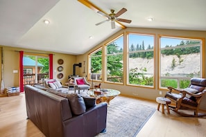 A spacious living room with a large sofa, a coffee table, and a rocking chair. There are large windows offering a view of a mountainous landscape.