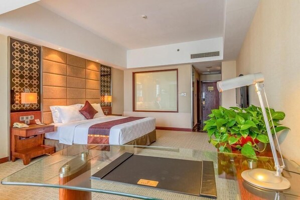 Una habitación de hotel con cama, mesitas de noche, una mesa de centro de vidrio, escritorio, silla, lámpara y planta en maceta.