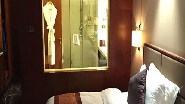 Een hotelkamer met een bed, een raam met uitzicht op een badkamer en een nachtkastje met een lamp.