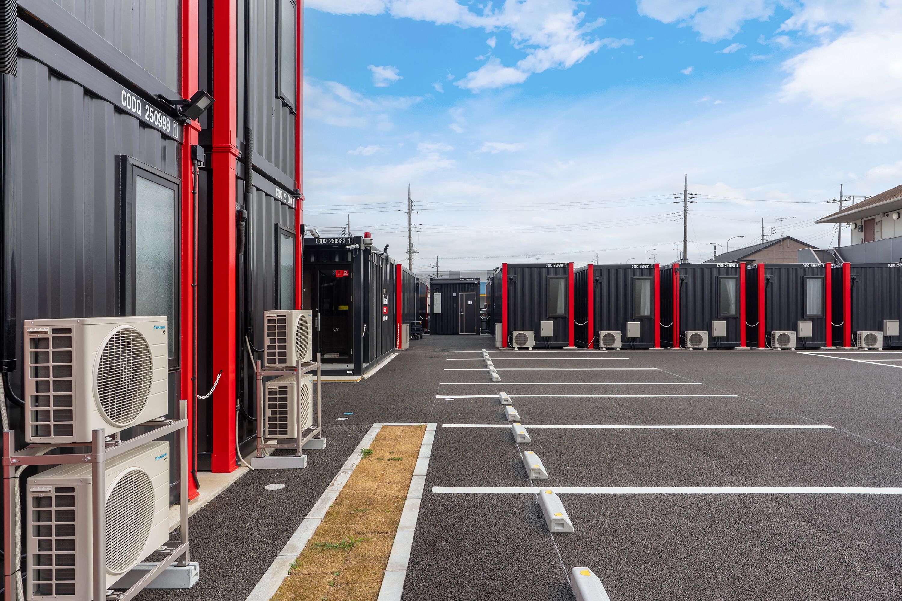A parking lot with rows of shipping containers, air conditioning units, and a building with red accents.