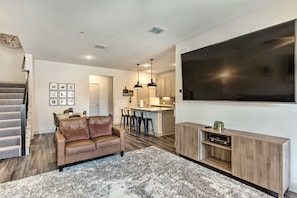 A modern living room with a sofa, a TV, and a kitchen area in the background. - Luxury Frisco Townhome Near The Star & PGA (Frisco)