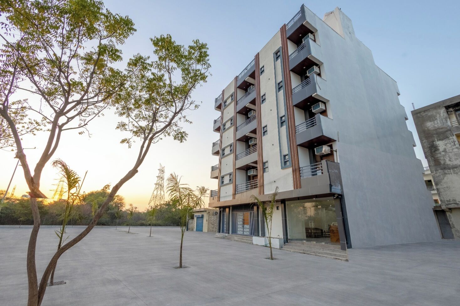A modern multi-story building with balconies and air conditioning units, surrounded by a concrete courtyard with sparse trees.