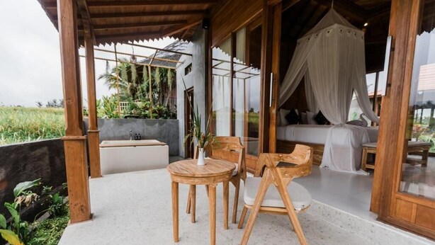 Un espacio exterior con asientos de madera, una mesa y sillas, un banco acolchado y una cama con dosel con ropa de cama blanca.