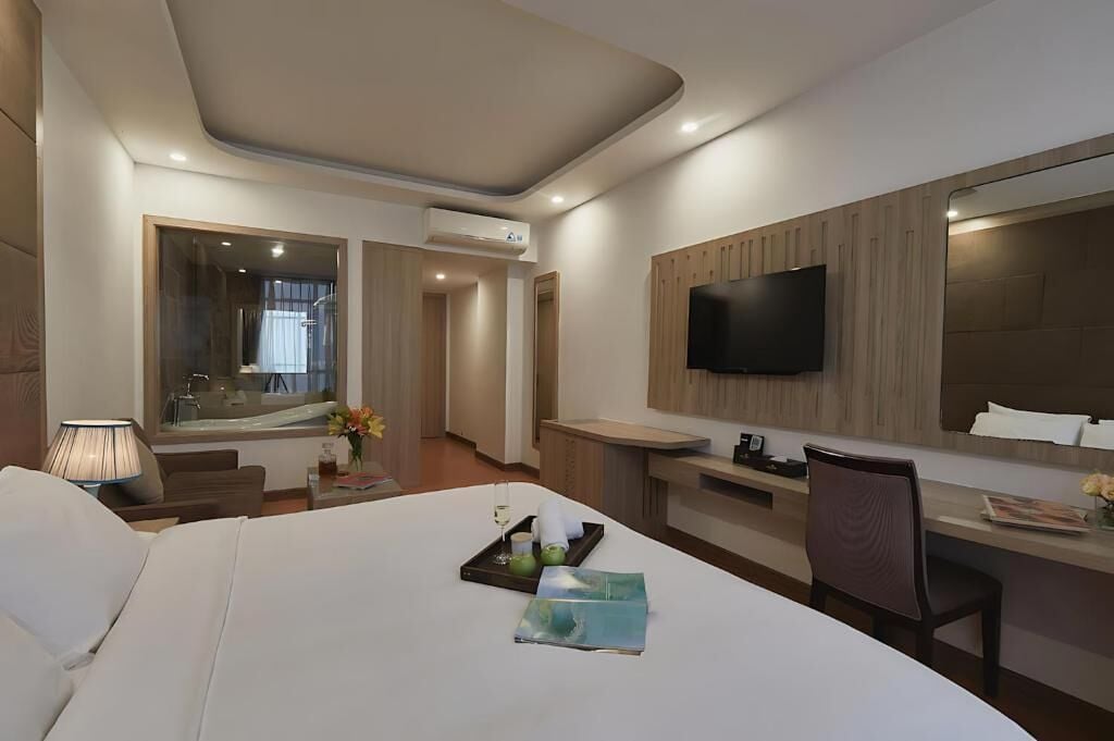 A modern hotel room with a large bed, a desk, a TV, and a bathroom visible through a glass door.