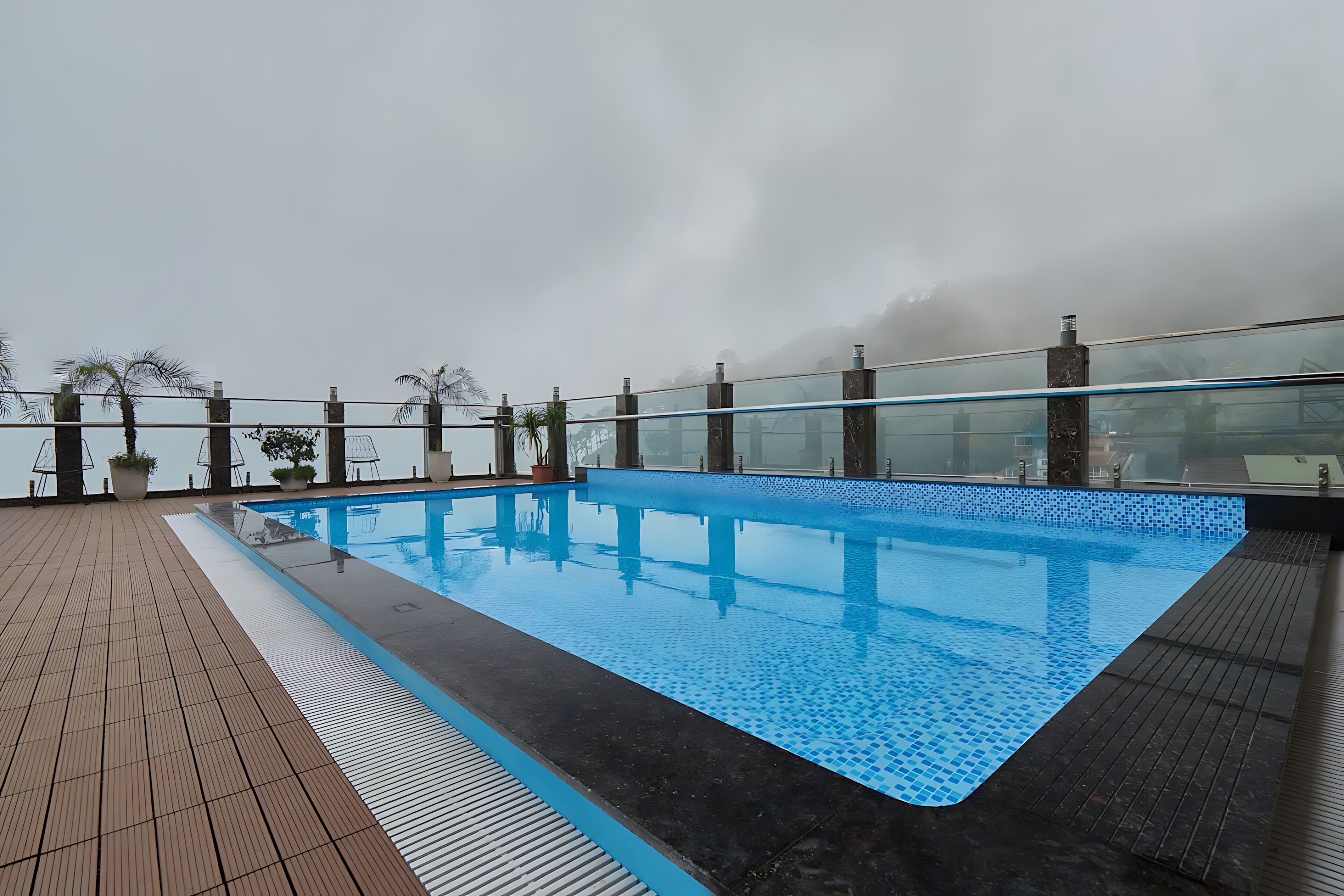 A rooftop pool with a clear blue water surface, surrounded by a wooden deck and glass railing.
