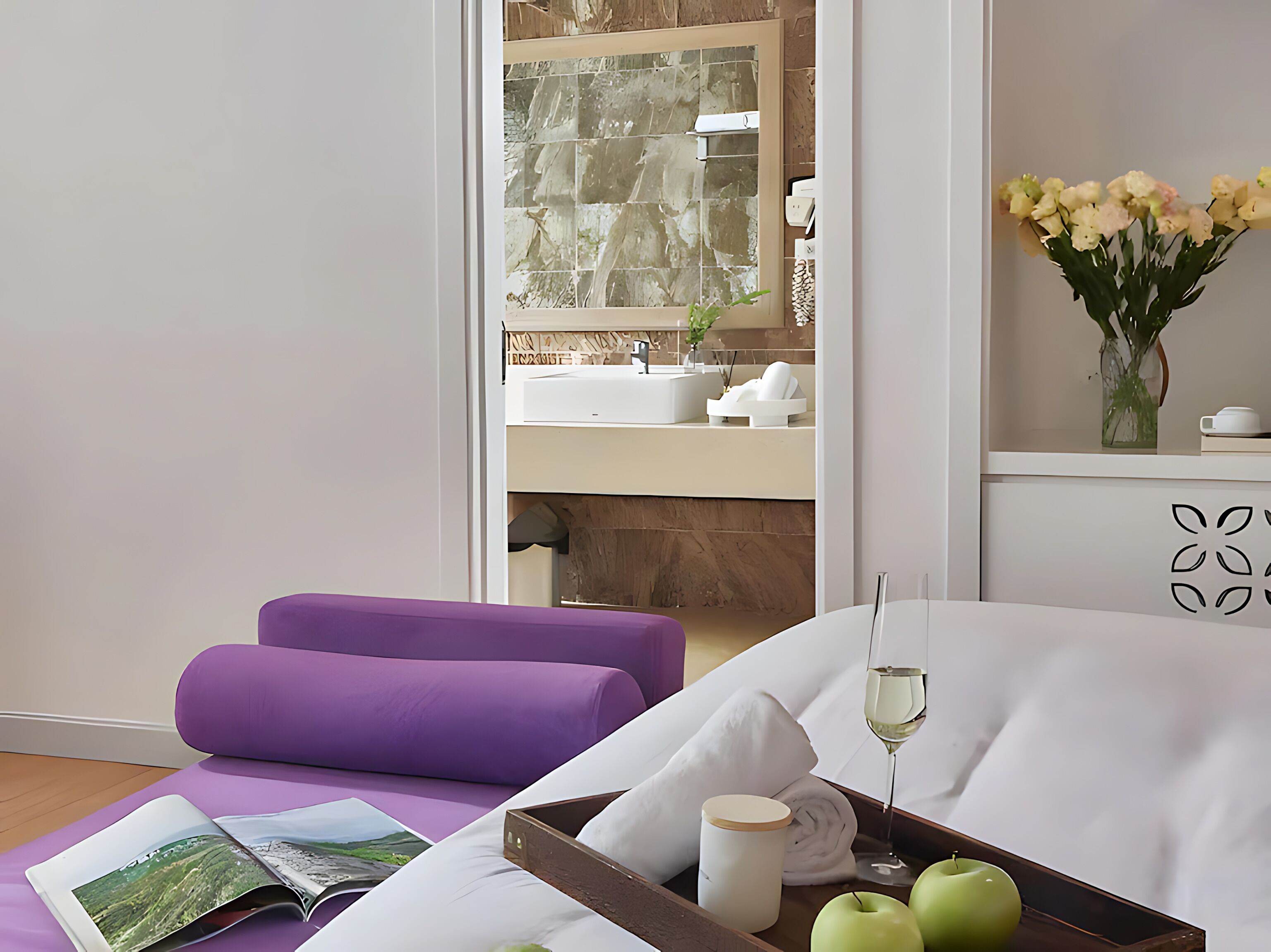 A hotel room with a bed, a purple chair, a tray with apples, a glass of wine, and a view of the bathroom.