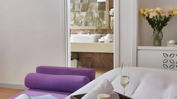 A hotel room with a bed, a purple chair, a tray with apples, a glass of wine, and a view of the bathroom.