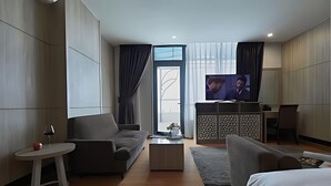 A modern hotel room with a sofa, a small table with a glass of wine, a television, and a large window with curtains.