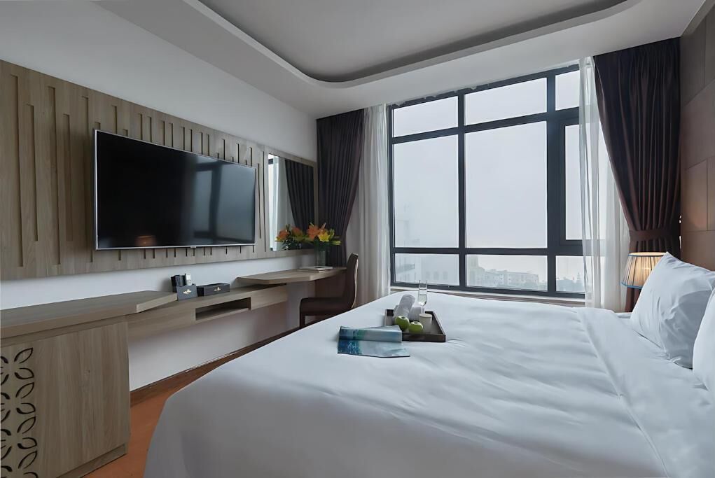 A hotel room with a large bed, a flat-screen TV mounted on the wall, a desk with a chair, and a large window offering a city view.