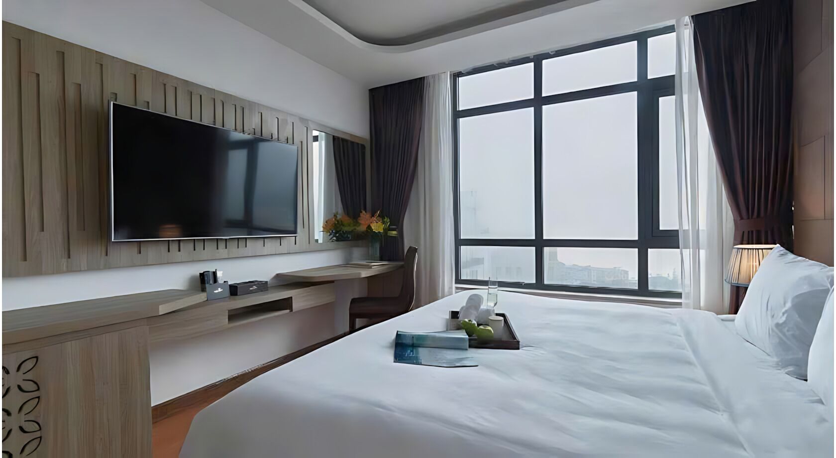Une chambre d’hôtel dotée d’un grand lit, d’un téléviseur à écran plat fixé au mur, d’un bureau avec une chaise et d’une grande fenêtre donnant sur la ville.