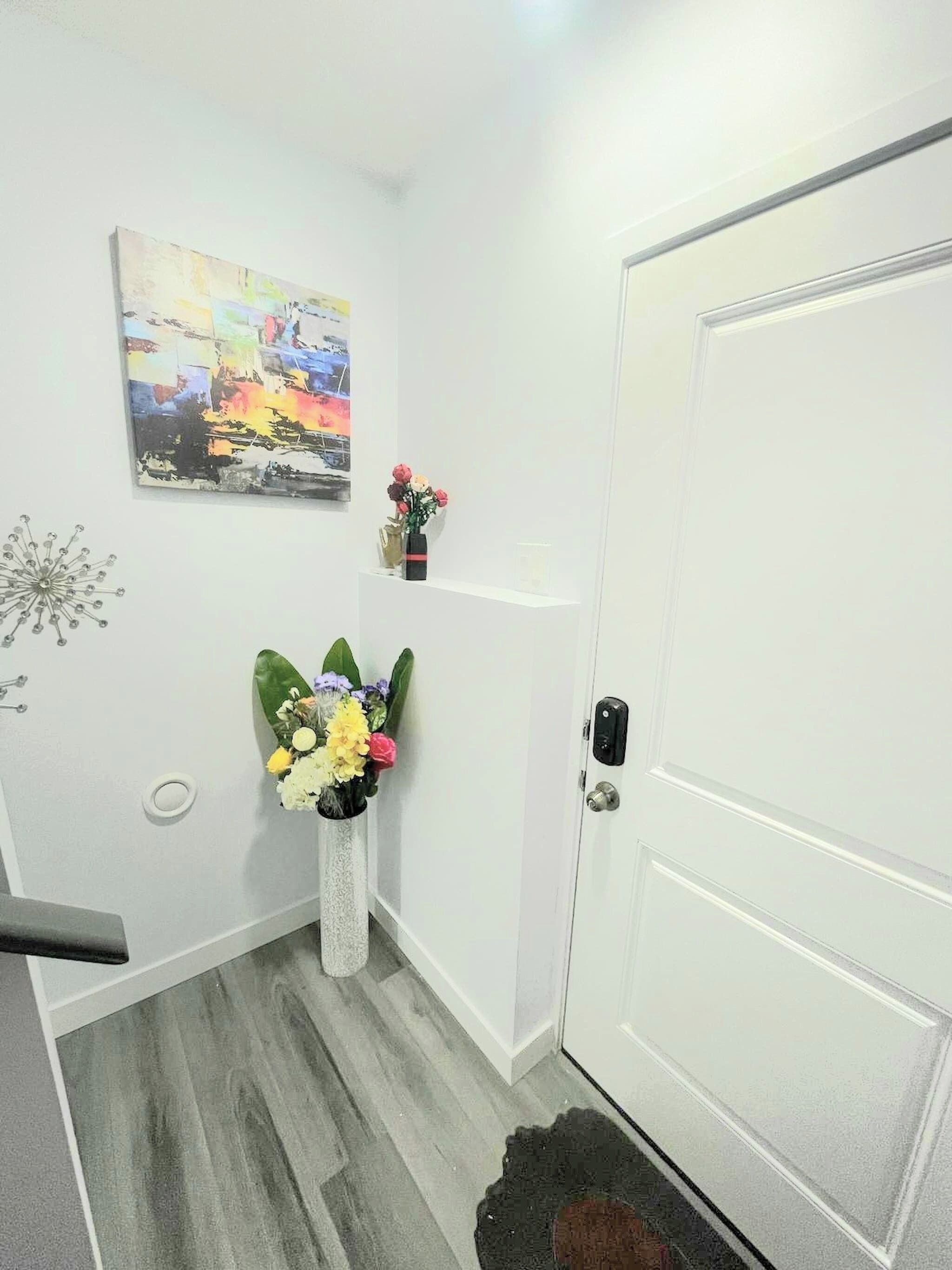 Un couloir avec une porte blanche, un tableau accroché au mur et un vase rempli de fleurs.