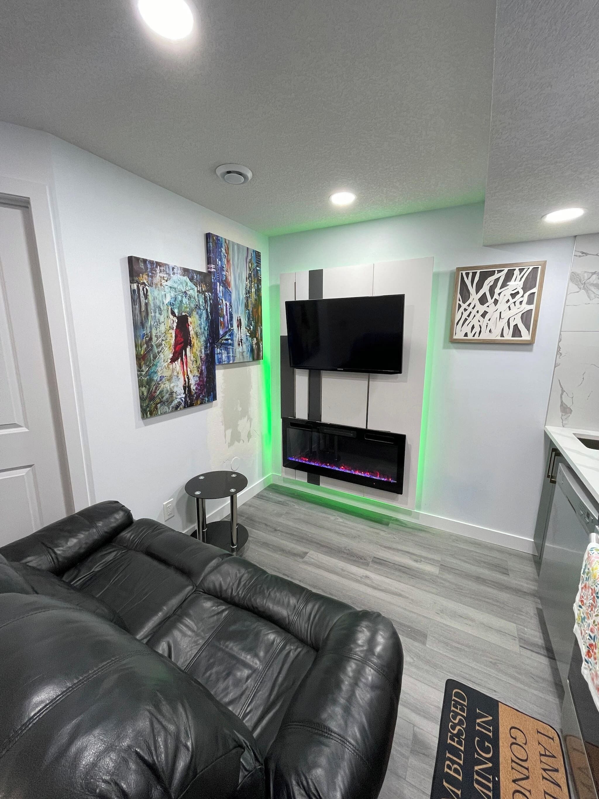 Un salon moderne avec un canapé en cuir noir, un téléviseur à écran plat fixé à un mur blanc et une cheminée décorative illuminée d’une lumière verte.