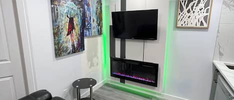Un salon moderne avec un canapé en cuir noir, un téléviseur à écran plat fixé à un mur blanc et une cheminée décorative illuminée d’une lumière verte.