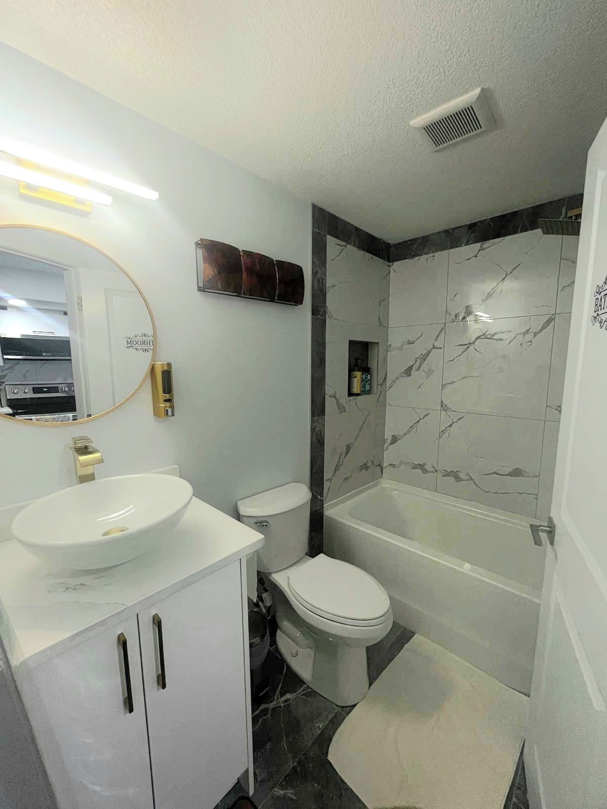 A modern bathroom with a white sink, toilet, and bathtub. There is a vanity with cabinets and a mirror. The walls are tiled with a marble pattern.