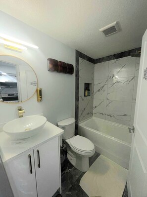 A modern bathroom with a white sink, toilet, and bathtub. There is a vanity with cabinets and a mirror. The walls are tiled with a marble pattern. - A Place of Refuge and Comfort (Edmonton)