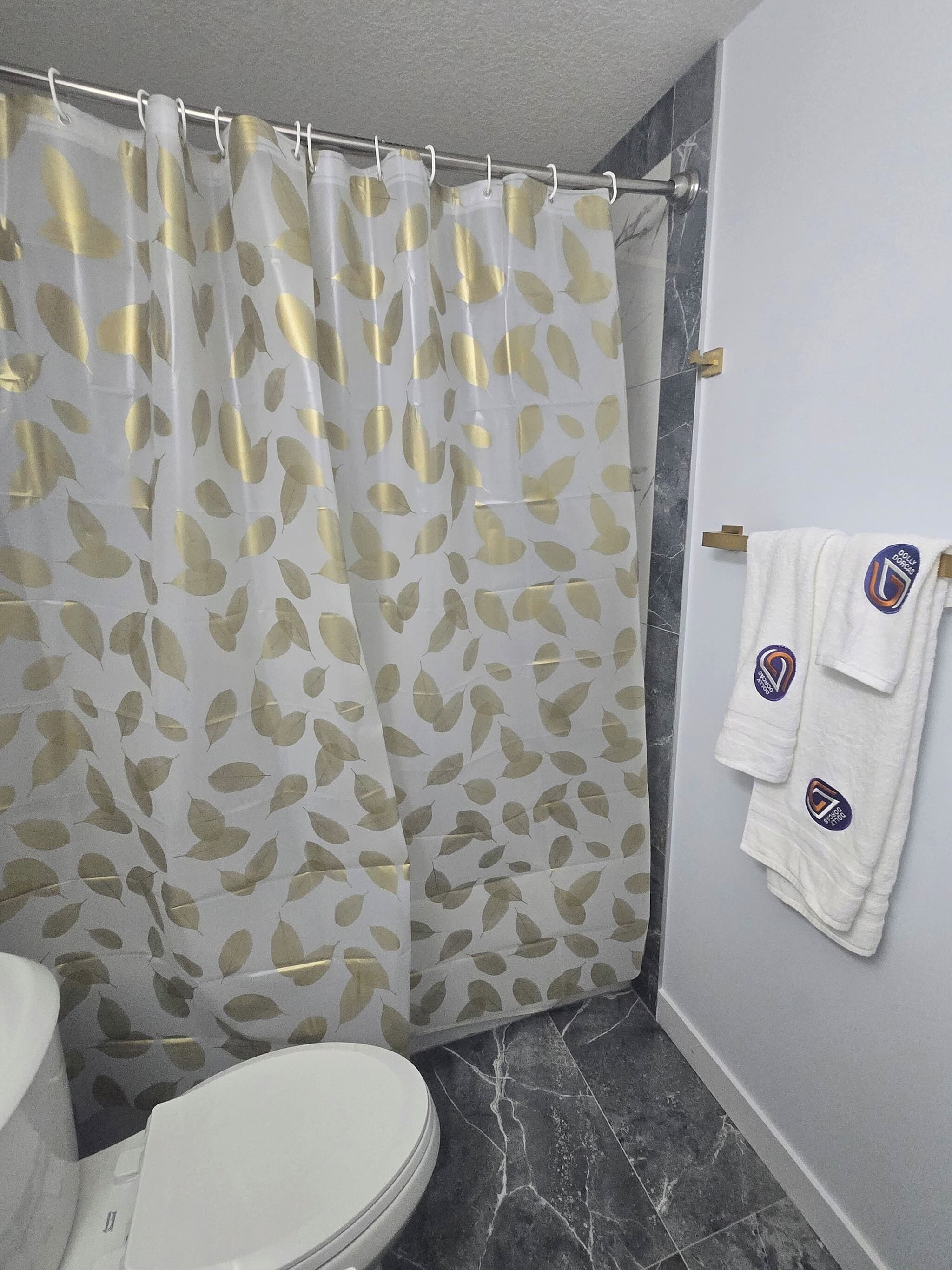 Une salle de bain avec un rideau de douche orné d’un motif de feuilles, des toilettes et deux serviettes avec un logo accroché au mur.