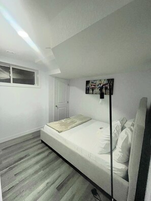 A bedroom with a single bed, a window, a door, and a painting on the wall. - A Place of Refuge and Comfort (Edmonton)