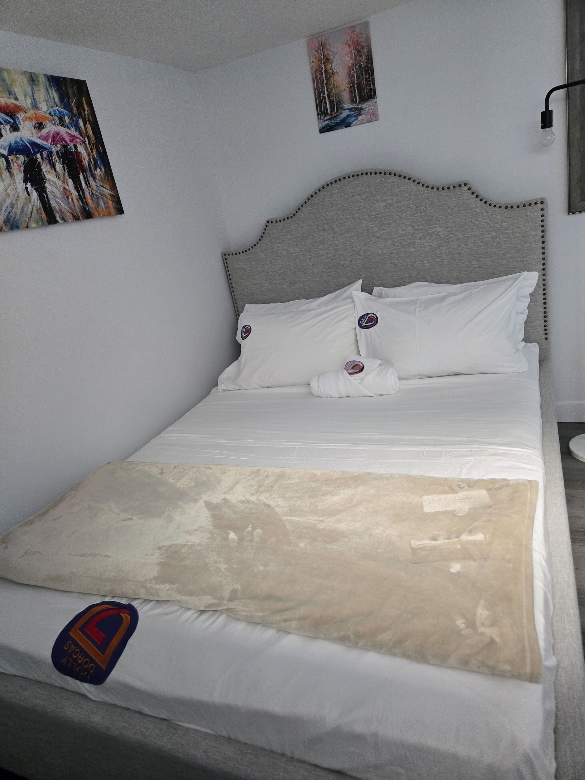 Ein ordentlich bezogenes Bett mit weißen Laken und einer beigen Tagesdecke, ein Kopfteil mit einem dekorativen Muster und zwei an der Wand hängende Kunstwerke, die Naturdarstellungen zeigen.