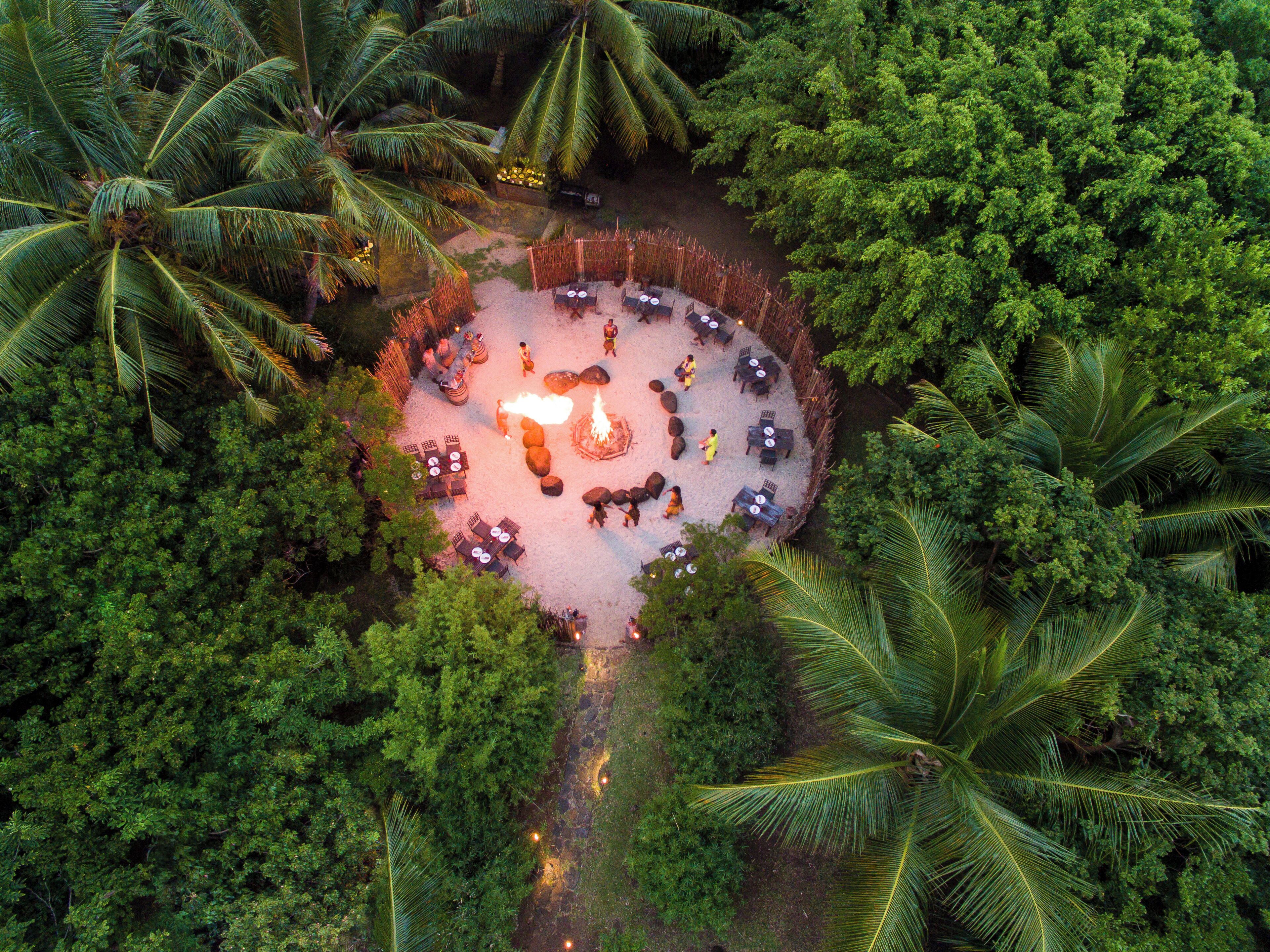 An aerial view of a circular outdoor seating area with tables and chairs, surrounded by dense tropical foliage.