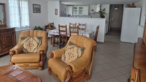A living room with two leather armchairs, a wooden coffee table, and a dining area with a table and chairs. A kitchen is visible in the background with a refrigerator and cabinets. - The gîte de la muraille welcomes you in the heart of nature, pets not allowed. (Mérignac)