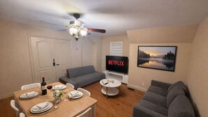 A living room with a dining table set for two, a grey sofa, a ceiling fan, a TV displaying 'NETFLIX', and a framed landscape painting. - Above-Ground · Sun-Filled · Not a Basement · Your Own Private Entrance (Brampton)