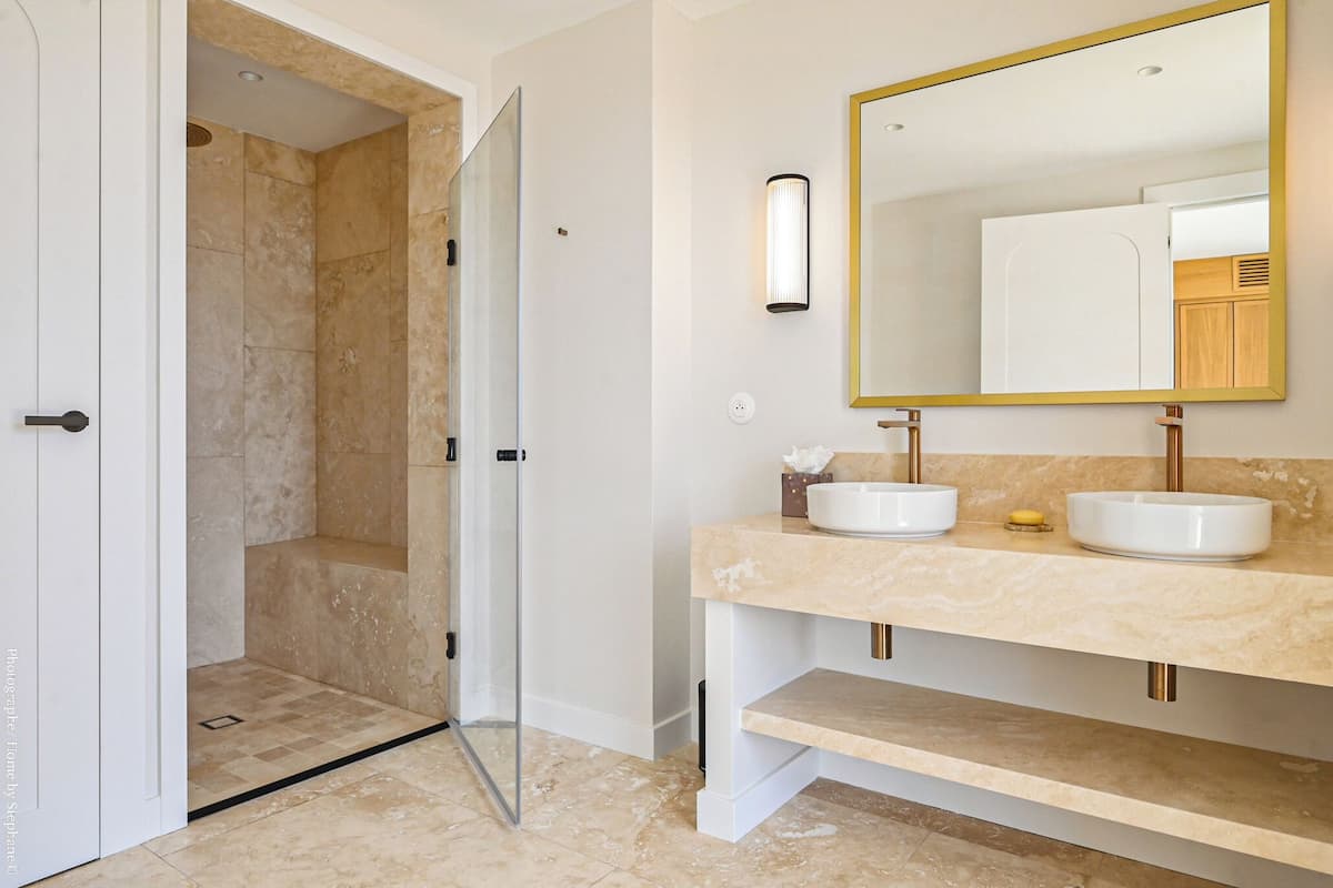 A modern bathroom with a glass-enclosed shower, two sinks with gold fixtures, and a large mirror with a gold frame.