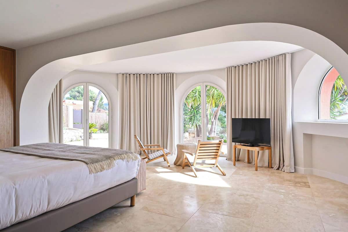 A bedroom with a bed, a TV, a seating area with large windows, and a view of greenery.