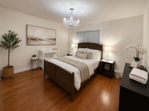 A bedroom with a wooden bed, bedside tables, a chandelier, a large window with curtains, a plant, and a welcome sign. - Poolside 4BR by Canterra Stays · Sleeps 12 (Brampton)