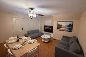 A living room with a dining area, a grey sofa, a TV displaying 'NETFLIX', and a painting of a lake. - 4BR · 2 Suites by Canterra Stays · Sleeps 8 (Brampton)