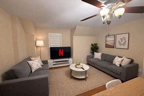 A living room with two grey sofas, a coffee table, a TV with a Netflix logo, a ceiling fan, and framed artwork on the wall. - 4BR · 2 Suites by Canterra Stays · Sleeps 8 (Brampton)
