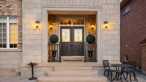 A building entrance with a dark double door, two round topiary decorations, and a small outdoor seating area with two chairs and a table. - Two Independent Suites · Two Entrances · Sleeps 8 · Fireplace + Sun-Filled Upper Unit (Brampton)