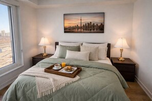 A hotel room with a bed, two bedside lamps, a tray with breakfast, and a cityscape painting on the wall. - Parking Included · Sauna & Climbing Wall · Steps to Square One · Weekly Stays (Mississauga)