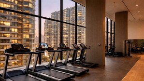 Une salle de sport équipée de tapis de course et d’appareils d’exercice, dotée de grandes fenêtres et offrant une vue sur les immeubles de grande hauteur.
