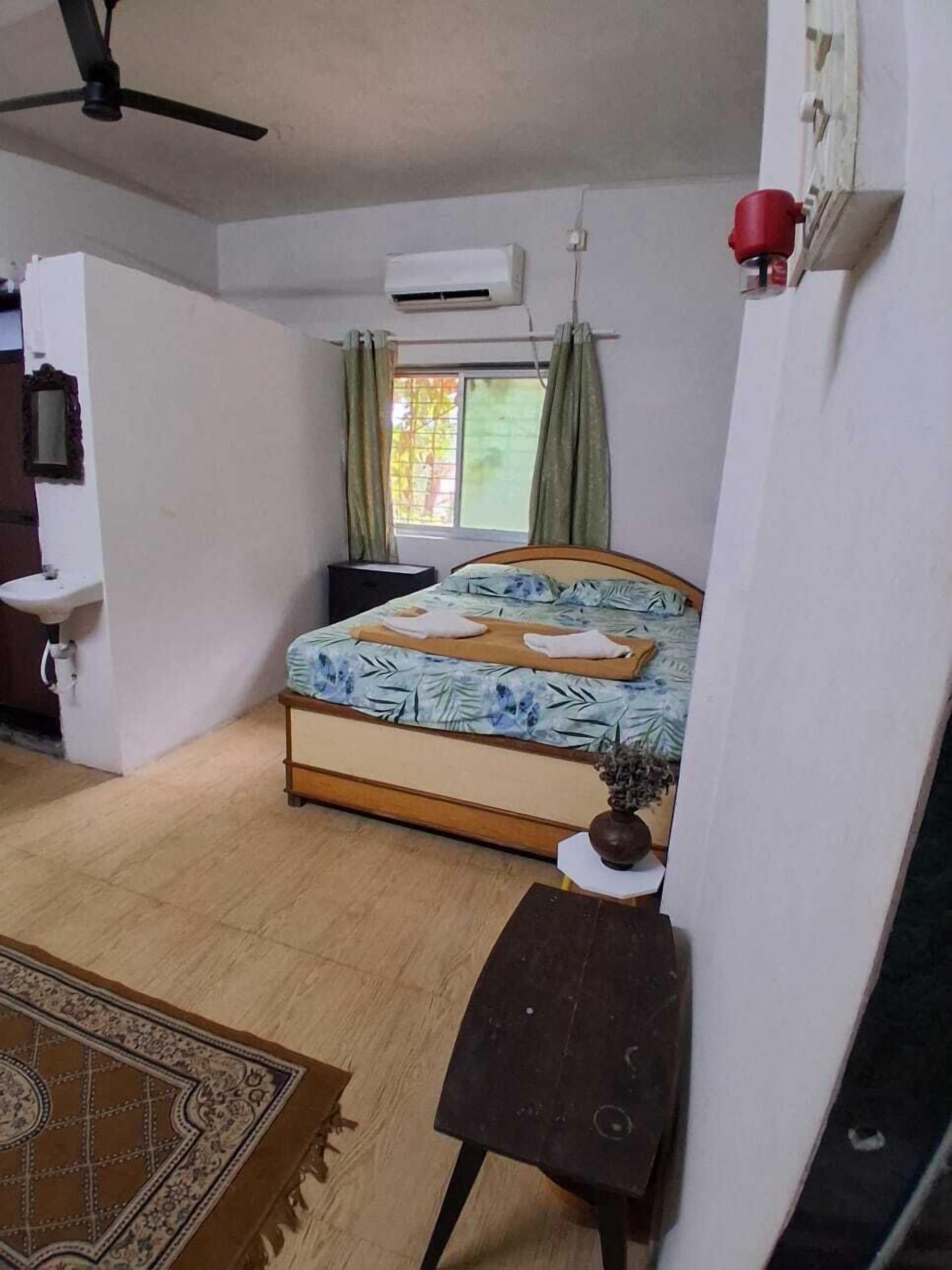 A bedroom with a bed, an air conditioner, a bedside table with a vase, a ceiling fan, and a window with curtains.