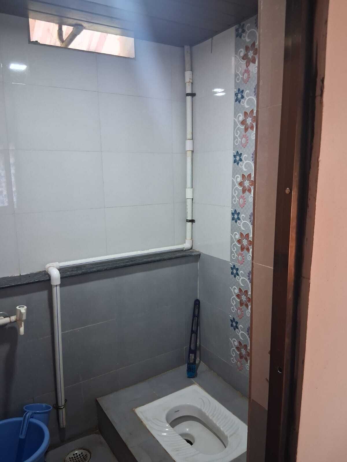 Een kleine badkamer met toilet, een blauwe emmer, een dweil en een raam met uitzicht op buiten.