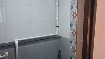 Een kleine badkamer met toilet, een blauwe emmer, een dweil en een raam met uitzicht op buiten.