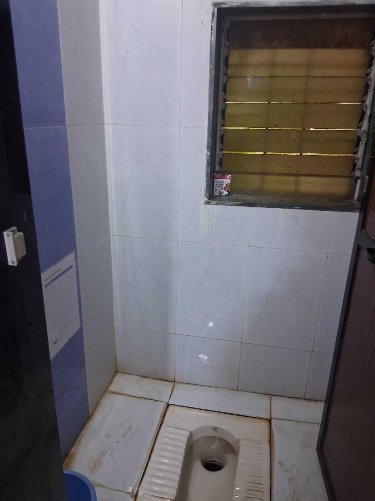 Een kleine betegelde badkamer met een toilet, een raam met tralies en een blauwe emmer.