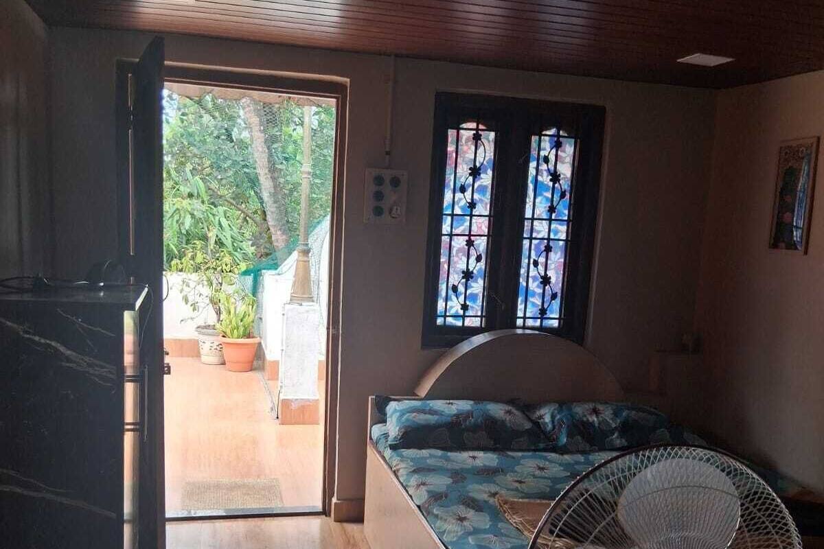 Una habitación con cama, ventilador, ventana con vista a follaje y puerta que da al exterior.