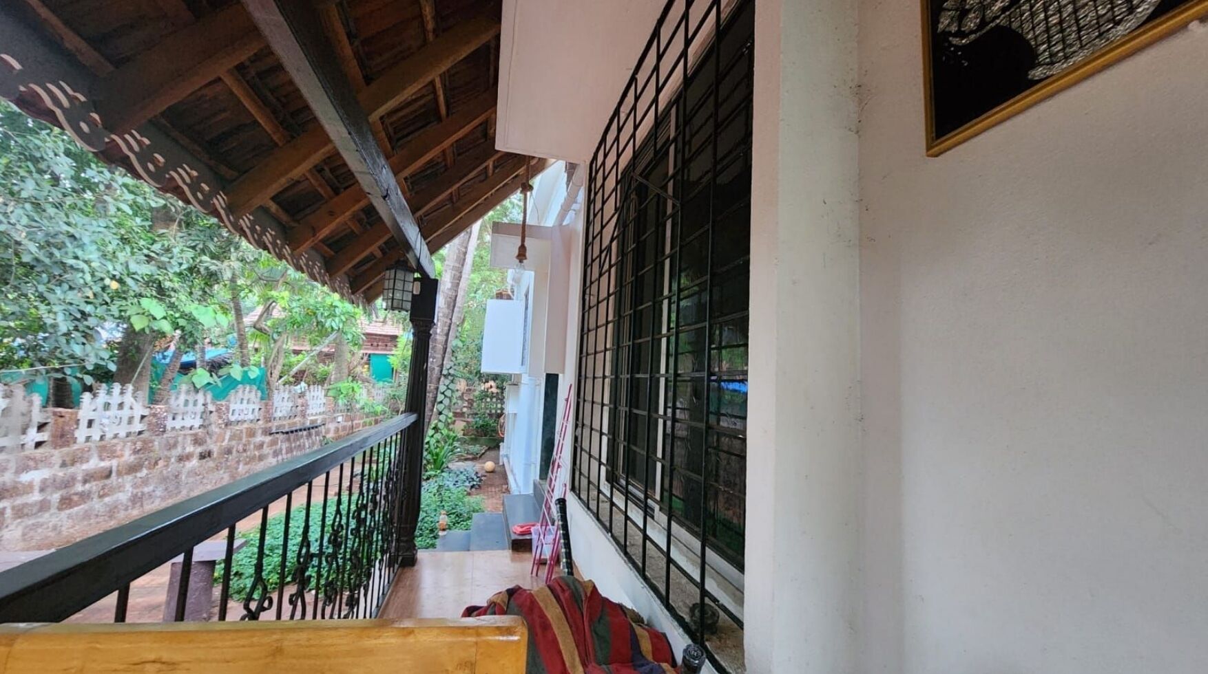 A balcony with a wooden railing, a view of a garden, and a window with metal bars.
