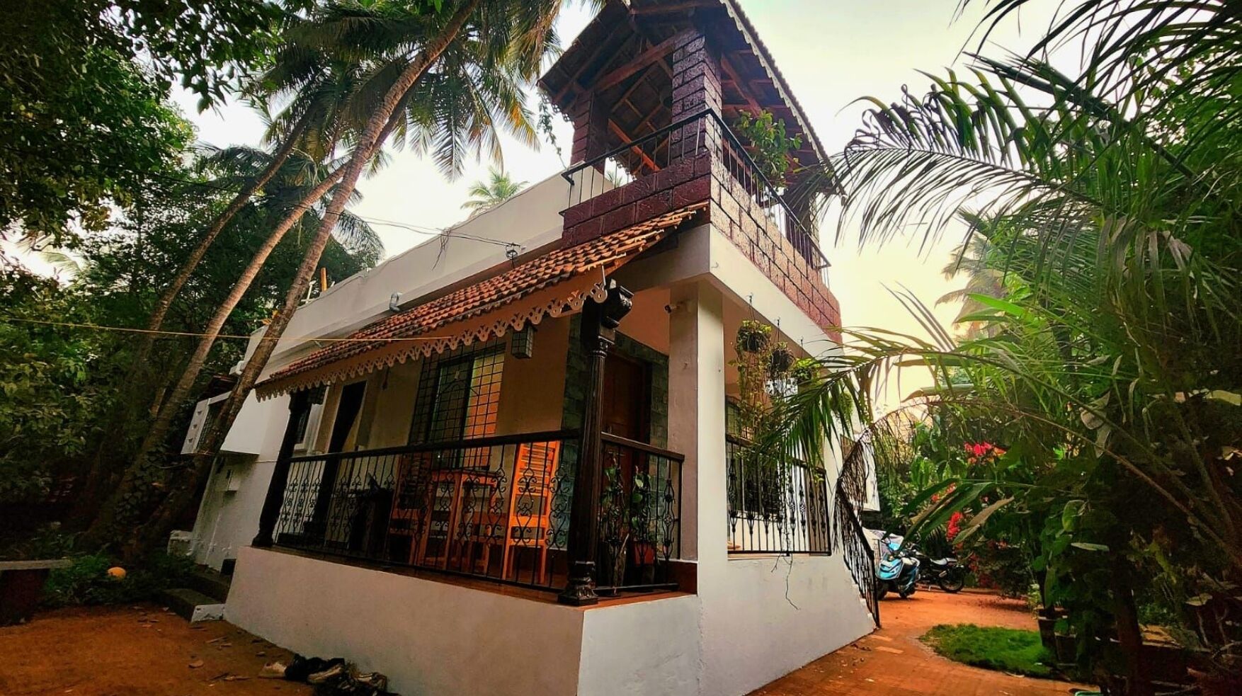 Une maison à deux étages dotée d’un balcon, entourée d’une végétation luxuriante et de palmiers.
