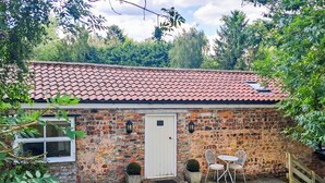 Een bakstenen huis met een pannendak, een witte deur en een klein terras met twee witte stoelen en een tafel.
