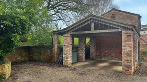 Een bogenpaviljoen van baksteen met een overdekte ingang, omgeven door bomen en een bakstenen muur.