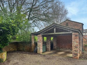 Een bogenpaviljoen van baksteen met een overdekte ingang, omgeven door bomen en een bakstenen muur.