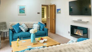 A living room with a blue sofa, a coffee table with a vase and fruit, a fireplace, and a television on the wall. - 2 bedroom accommodation in Rock (Rock)