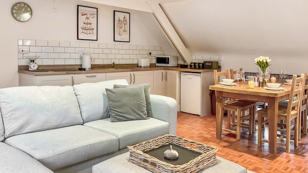 Een modern loft appartement met een zithoek, bestaande uit een lichtgrijze bank, een houten eettafel voor vier personen, en een keuken met witte apparatuur en een witte betegelde achterwand.