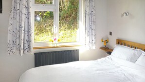 Ein ordentlich bezogenes Bett mit einem Buch darauf, ein Fenster mit Vorhängen, ein Nachttisch mit Blumen in einer Vase und ein Heizkörper unter dem Fenster.