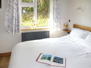 Un lit bien fait, avec un livre posé dessus, une fenêtre avec des rideaux, une table de chevet ornée d’un vase de fleurs et un radiateur placé sous la fenêtre.
