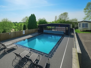 A rectangular outdoor swimming pool with a covered seating area, surrounded by a wooden fence and a grassy area with trees.