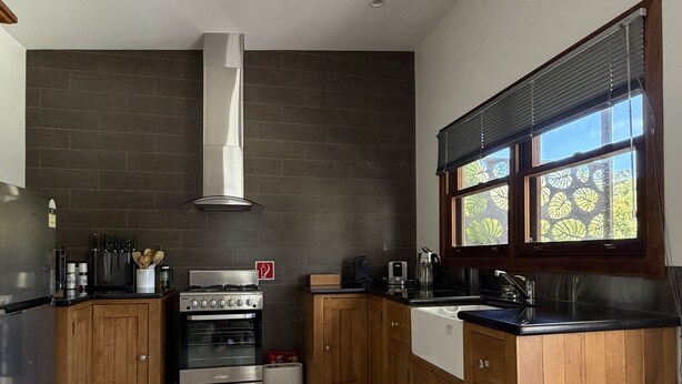 A modern kitchen with dark tiled walls, wooden cabinets, a stainless steel stove, and a window with blinds. - Cabin One- Sauna, pet-friendly, adults only (Lowanna)